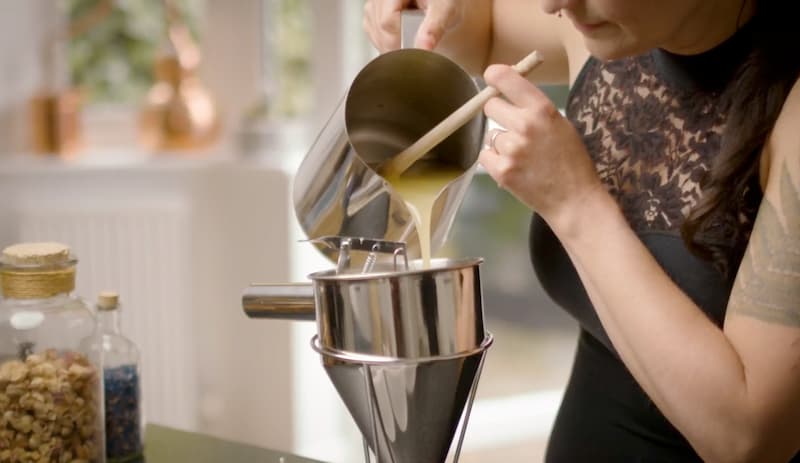 A woman carefully pours molten wax from a stainless steel jug into a wax dispenser for precise candle measuring