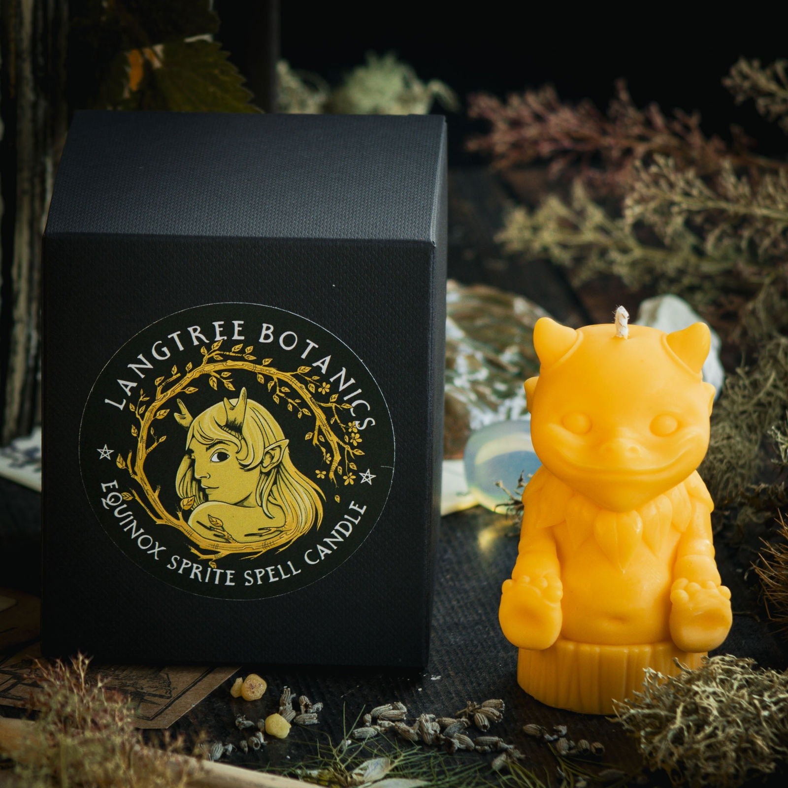 A bright yellow candle in the shape of a mythical nature sprite sits next to a black cardboard packaging box