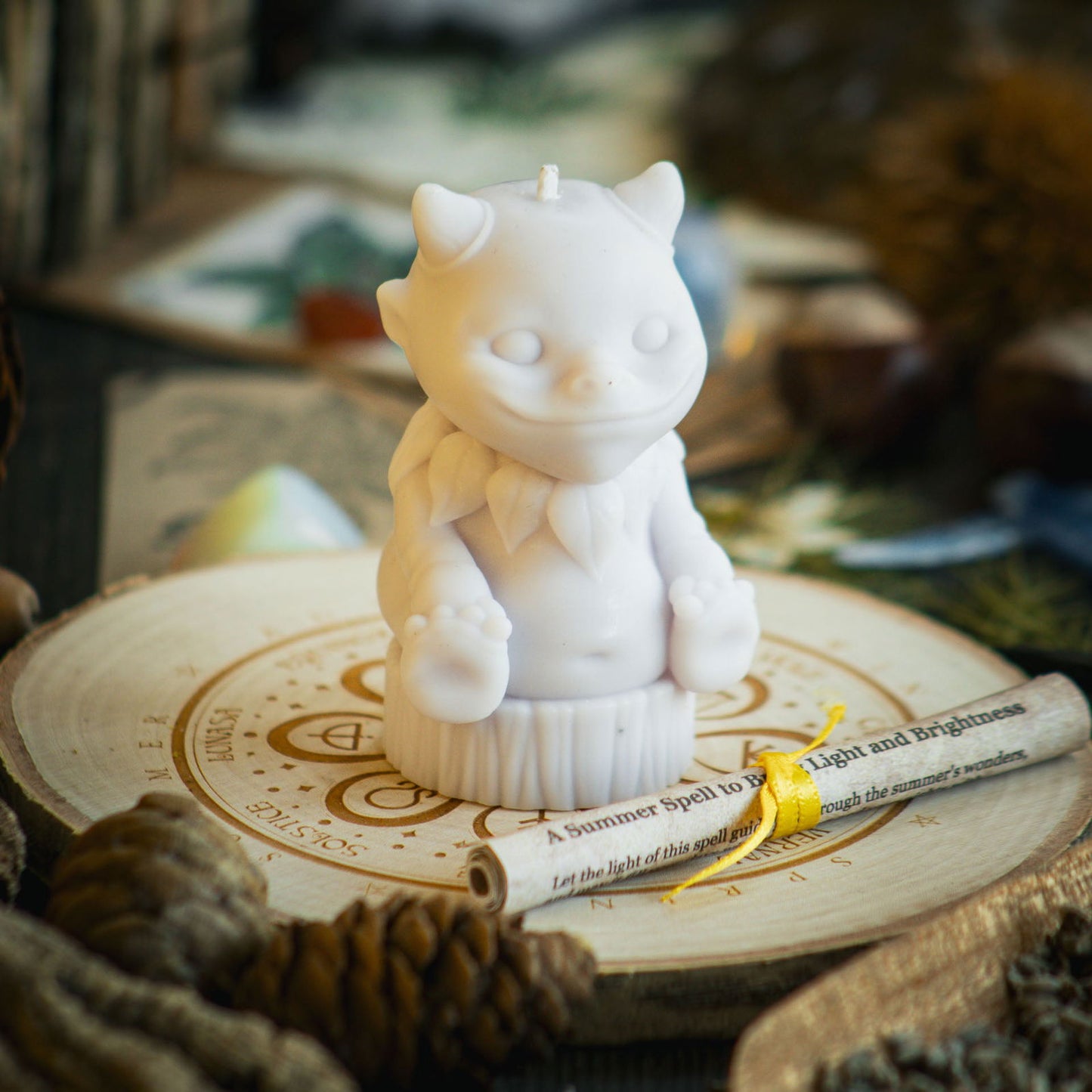 A pale lavender candle in the shape of a magical nature sprite sits behind a rolled up spell to celebrate the summer solstice