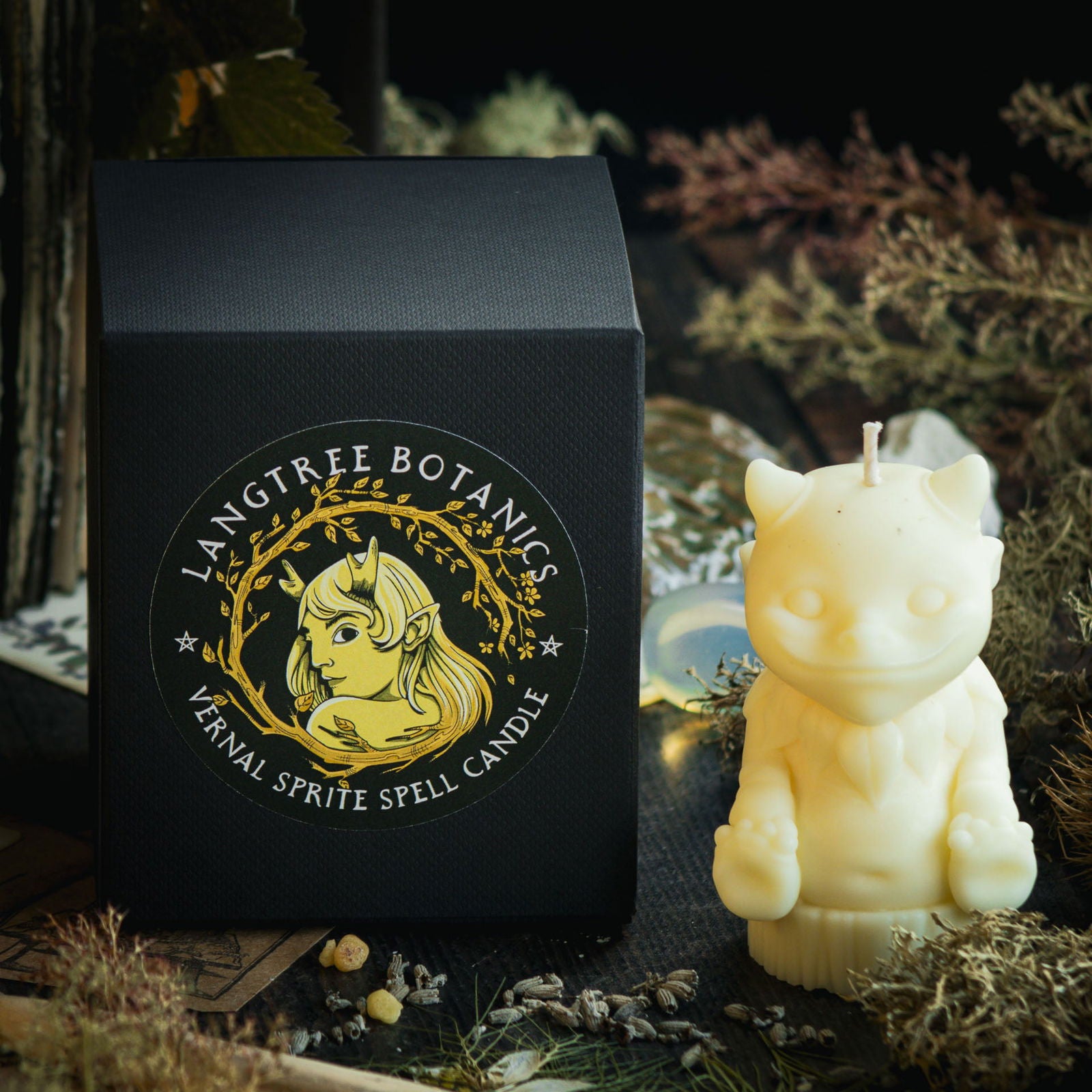 A light pale yellow candle in the shape of a mythical nature sprite sits next to a black cardboard packaging box