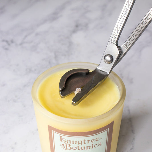 A silver stainless steel candle wick trimmer trims the top of a yellow candle on a marble background