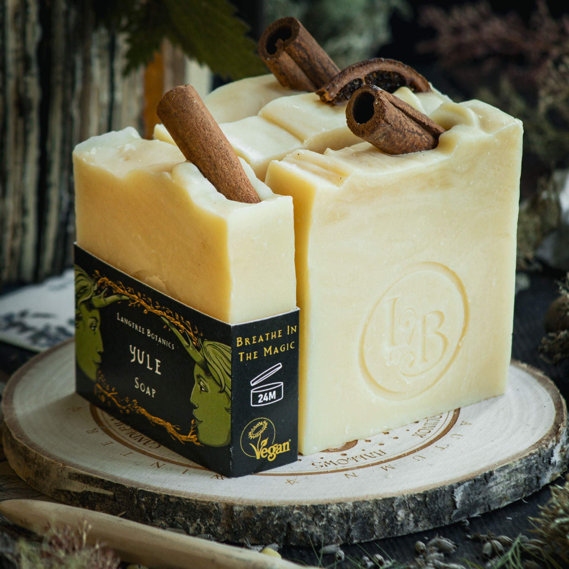 Four bars of Yule soap on top of a decorative wheel-of-the-year log slice surrounded by seasonal foliage.