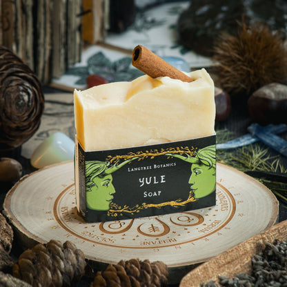 A bar of Yule soap on top of a decorative wheel-of-the-year log slice surrounded by seasonal foliage